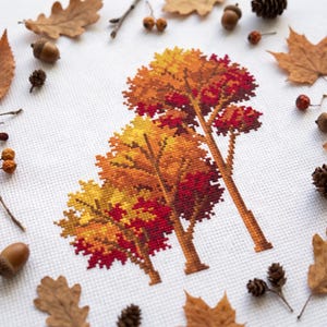 Pode incluir: Um bordado em ponto cruz sobre tecido branco apresentando duas árvores com folhagem de outono em tons de amarelo, laranja e vermelho profundo. As árvores têm troncos e galhos castanhos, com as folhas em um padrão de grade pixelado.