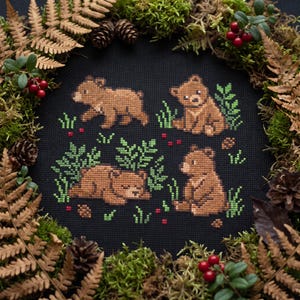 May include: A black fabric square featuring a cross-stitch pattern of four brown bear cubs in various poses, surrounded by green foliage and small red berries. The bears are depicted walking, sitting, and lying down among stitched plants.