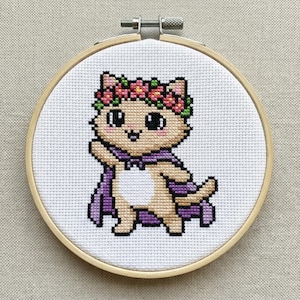 May include: A cross-stitch embroidery hoop featuring a cartoon cat wearing a purple cape and a floral crown. The cat is tan with black eyes and pink cheeks. The embroidery is set in a wooden hoop.