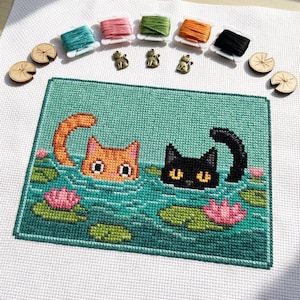 Op de afbeelding: Kruissteekborduurwerk van twee katten in een vijver met waterlelies en roze bloemen. De katten zijn oranje en zwart. Boven de borduursel bevinden zich spoelen garen, kikkerbedels en houten waterlelieknoppen.
