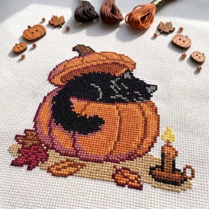 May include: A cross-stitch embroidery on white fabric featuring a black cat curled inside a large orange pumpkin. The design includes a small lit candle, scattered autumn leaves, and wooden pumpkin-shaped buttons and embroidery floss.