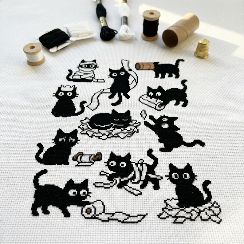 May include: A cross-stitch pattern featuring black cat designs in various poses, including wrapped in bandages, playing with toilet paper, and lounging. The image also shows spools of thread, a thimble, and a cardboard tube.
