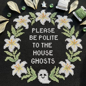 Może przedstawiać: Czarny haft krzyżykowy z napisem "PLEASE BE POLITE TO THE HOUSE GHOSTS" otoczony kwiatowym wieńcem. Wieniec zawiera białe kwiaty z żółtymi środkami i zielonymi liśćmi. Mały duch jest wyszyty na dole.