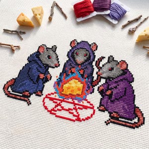 May include: A cross-stitch embroidery on white fabric featuring three gray mice wearing hooded robes in blue, purple, and dark purple. The mice are gathered around a central fire with orange and yellow flames atop a red circular pentagram design.
