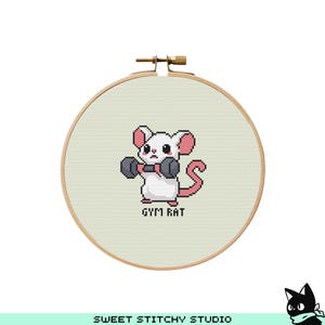 Op de afbeelding: Een witte muis met roze oren en een zwarte neus tilt een halter. De muis bevindt zich op een beige achtergrond met de tekst "Gym Rat" eronder. De afbeelding is in een ronde houten borduurring.