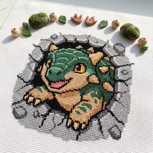 Könnte beinhalten: Eine Kreuzstichstickerei auf weißem Stoff, die einen grünen und beigen Baby-Dinosaurier zeigt, der aus einer rissigen grauen Steinmauer hervorbricht. Das Design ist von kleinen dekorativen Elementen wie Moossteinen, Holzperlen und Blättern umgeben.