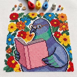 May include: A cross-stitch pattern on white fabric featuring a purple pigeon wearing round glasses and reading a pink book. The pigeon is surrounded by a vibrant border of red, yellow, and blue flowers. Red, yellow, and blue embroidery floss, wooden buttons, and a metal thimble are arranged at the top.