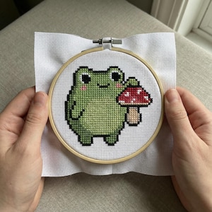 Puede incluir: Bordado de punto de cruz de una rana verde sosteniendo una seta roja y blanca. La rana tiene ojos negros y mejillas rosas. El bordado está en un aro de madera sobre tela blanca.