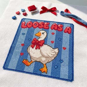 May include: A cross-stitch design featuring a white goose with a red bow, walking on a blue striped background. The text "LOOSE AS A" is in red. Small red hearts and blue heart shapes are scattered around the goose. Additional craft supplies are visible.