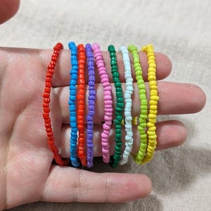 May include: A collection of nine thin bracelets made from small, round, multi-coloured glass seed beads. The bracelets feature individual strands in red, blue, orange, purple, pink, dark green, white, lime green, and bright yellow colours.
