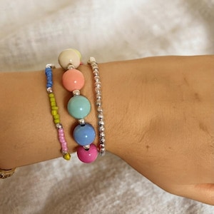 Puede incluir: Un conjunto de tres pulseras compuesto por una hilera de pequeñas cuentas de semillas multicolores, una hilera de grandes cuentas esféricas lisas en tonos pastel crema, melocotón, turquesa, azul y rosa, y una hilera de cuentas plateadas.