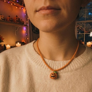 Peut inclure: Un collier composé d'une chaîne de petites perles orange et d'un pendentif citrouille d'Halloween orange avec une tige verte. Le pendentif présente un visage sculpté avec des yeux, un nez et un large sourire édenté noirs au centre.