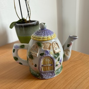 Peut inclure: Une théière décorative en céramique en forme de cottage. La théière présente un toit violet et jaune, une porte et des fenêtres. Elle est ornée de motifs floraux et d'une base verte. La poignée et le bec sont décorés de motifs floraux.