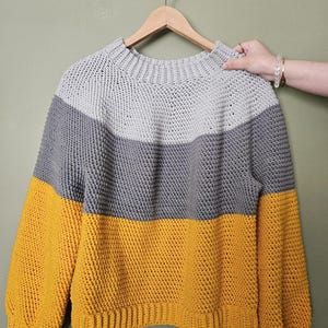 May include: A color-blocked sweater in gray, dark gray, and mustard yellow hangs on a wooden hanger. The sweater has a ribbed neckline and cuffs, with a textured knit pattern. The sleeves are a mustard yellow color.