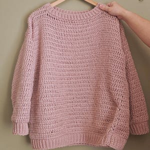 May include: A light pink, oversized crochet sweater with a crew neck and long sleeves. The sweater is hanging on a wooden hanger. The crochet stitch creates a textured appearance. The sleeves have ribbed cuffs.