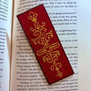 Puede incluir: Un marcapáginas rojo con un diseño bordado en oro. El diseño presenta un símbolo estilizado con el texto "Stay Safe" en oro.