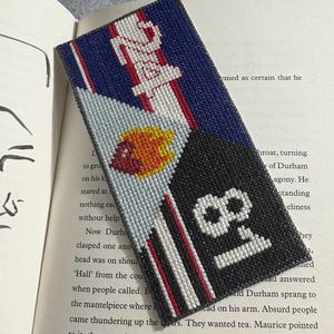 May include: Needlepoint bookmark with a design featuring the number "18" in white on a black background, a red and white striped design, and the word "24" in white on a blue background. The bookmark is resting on the pages of a book.