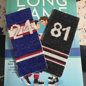 May include: Two beaded bookmarks rest on a book titled "The Long Game" by Rachel Reid. One bookmark is blue with the number 24, the other is black with the number 81. Both have red and white stripes. The book cover is teal with illustrations.