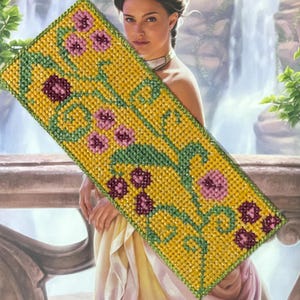 Puede incluir: Un marcapáginas amarillo con un diseño floral verde y rosa. El marcapáginas está hecho de tela de punto de cruz.