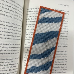 May include: A handmade bookmark with a white and blue wave pattern, framed by an orange border. The bookmark is made of stitched fabric and rests on the pages of a book.