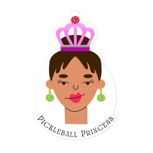 Puede incluir: Una pegatina con una ilustración de dibujos animados de una persona con cabello castaño, que lleva una corona rosa y pendientes de lunares verdes. El texto "PICKLEBALL PRINCESS" está impreso debajo.