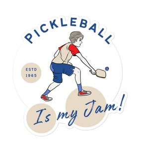 Puede incluir: Una pegatina con una ilustración de dibujos animados de una persona jugando al pickleball. El diseño incluye la palabra "PICKLEBALL" en azul, la frase "Is my Jam!" en escritura azul y "ESTD 1965". La persona viste ropa roja, azul y beige.