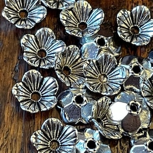May include: A collection of silver-colored, flower-shaped metal bead caps. Each cap has five petals with detailed lines radiating from a central hole. The beads are scattered on a dark wooden surface, creating a textured contrast.