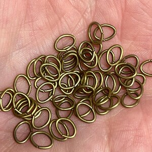 May include: A collection of small, oval-shaped brass jump rings. The rings are a warm, antique gold color and are scattered across a surface. These jewelry findings are ideal for crafting and jewelry making projects.