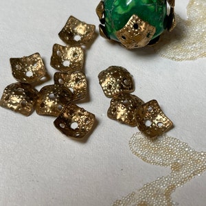 May include: A collection of ten gold-toned metal square-shaped embellishments with a textured surface and small holes. The embellishments are scattered on a white surface. A green and gold-toned decorative bead is also visible in the image.