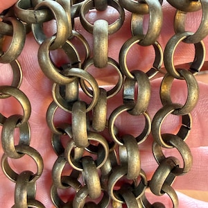 May include: Close-up of a section of a brass chain. The chain is composed of interlocking oval links, creating a textured surface. The metal has a slightly aged, antique appearance, with variations in color and tone.