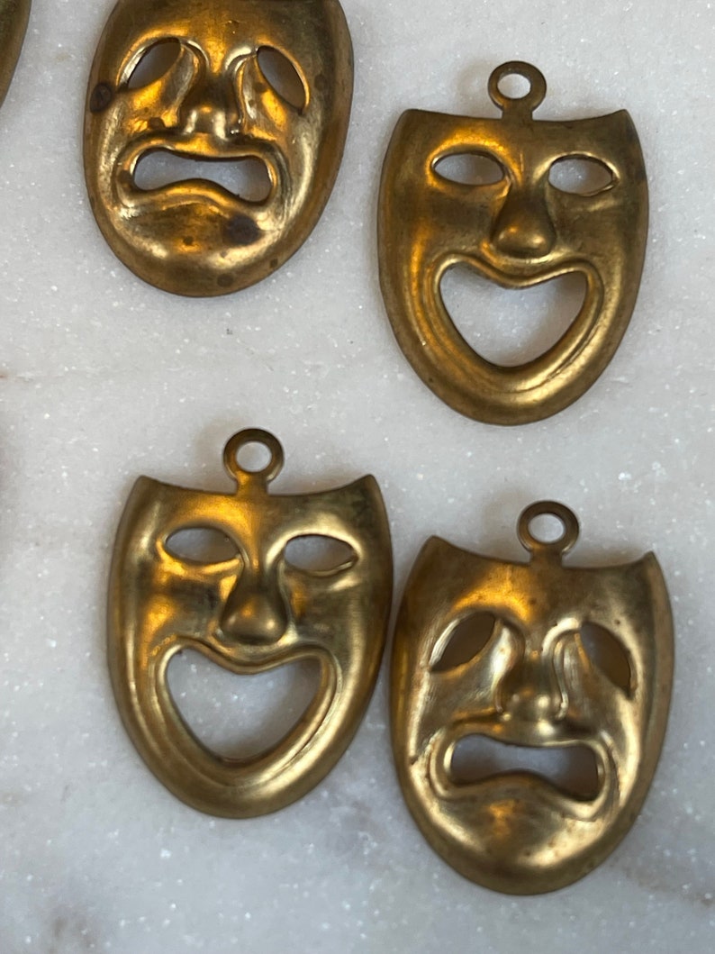 May include: Four gold-colored metal masks with a theatrical theme. Two masks depict a smiling face and two masks depict a frowning face.