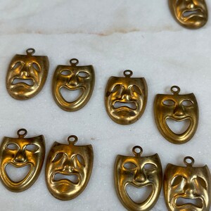 May include: Ten gold-colored metal mask charms with a theatrical theme. The masks have a variety of expressions, including happy, sad, and surprised.