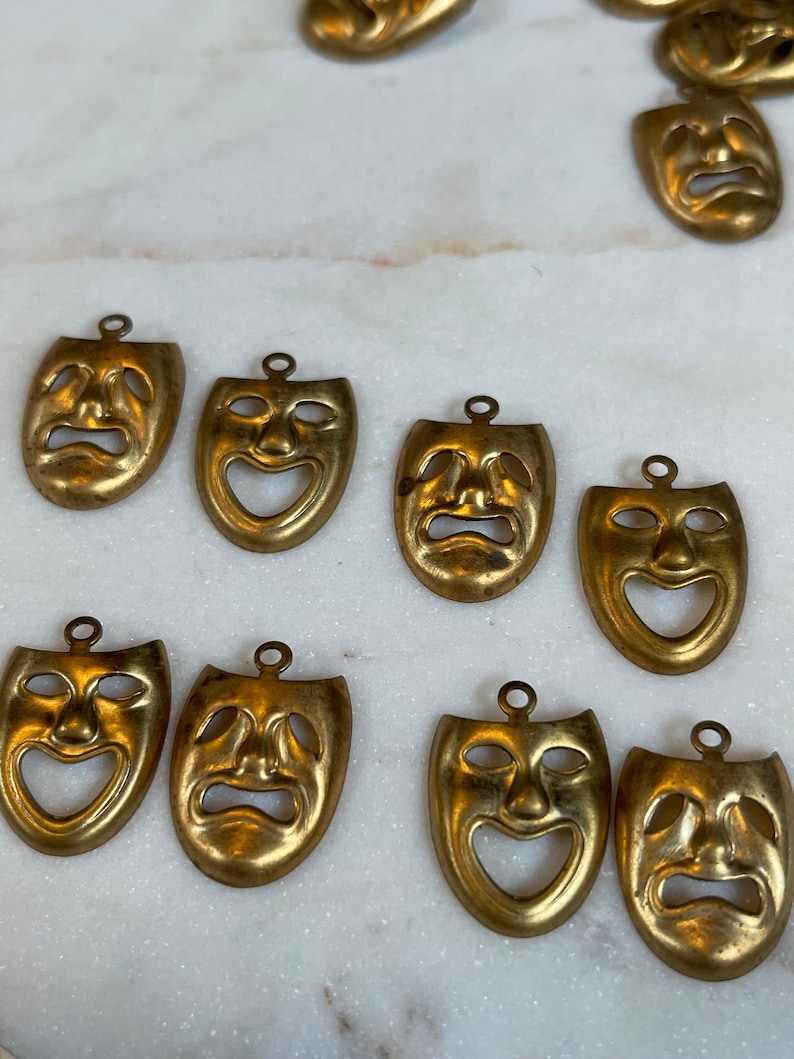 May include: Ten gold-colored metal mask charms with a theatrical theme. The masks have a variety of expressions, including happy, sad, and surprised.