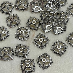 May include: Silver-toned metal square beads with an ornate, openwork design. Each bead features a central hole and swirling patterns, ideal for jewelry making or craft projects. The beads are scattered on a light-colored surface.