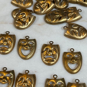May include: A collection of 18 gold-toned metal mask charms with a distressed finish. The masks have a classic theater design with a sad or happy expression.