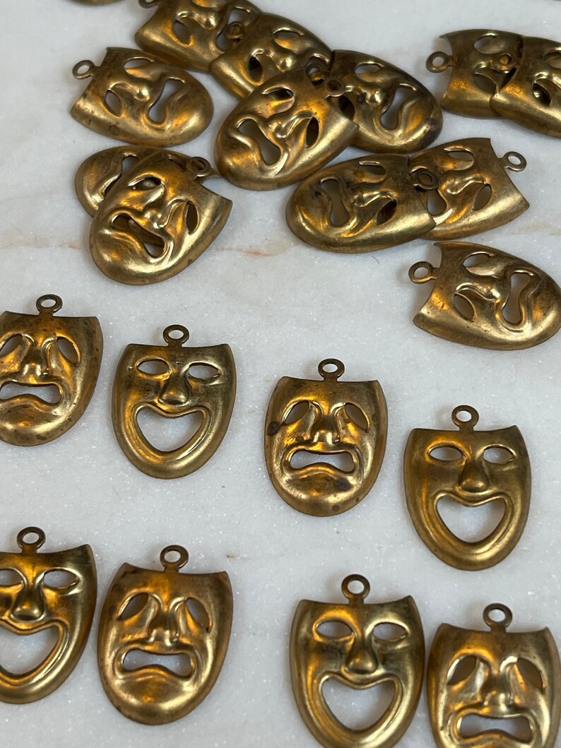 May include: A collection of 18 gold-toned metal mask charms with a distressed finish. The masks have a classic theater design with a sad or happy expression.