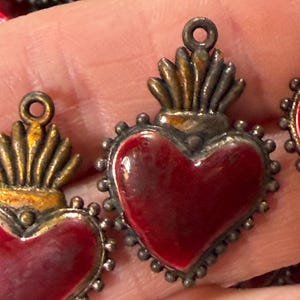 May include: A collection of heart-shaped charms featuring a deep red enamel center surrounded by a dark, oxidized silver-toned metal frame. Each charm is topped with a stylized flame motif and includes a small circular loop for attachment.