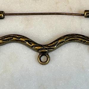 May include: A gold-toned metal safety pin with a decorative, ornate design. The pin has three loops on the bottom.
