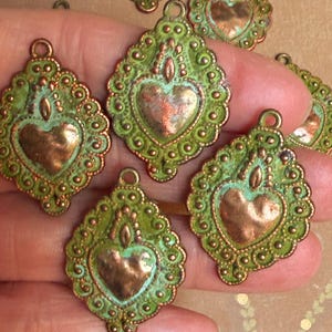 May include: Several ornate, heart-shaped pendants with a distressed, antique appearance. The pendants feature a copper-colored heart in the center, surrounded by a green, textured frame with a scalloped edge. Each pendant has a small loop at the top.