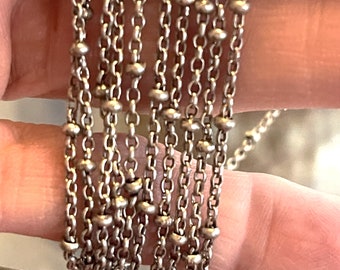 May include: Close-up of multiple strands of a silver-colored necklace. The necklace features a delicate chain interspersed with small, round beads. The chains are layered, creating a textured and intricate design, suitable for jewelry.