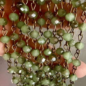 May include: A multi-strand necklace featuring numerous sage green faceted glass beads linked together with antique bronze-toned metal wire. The beads are arranged in vertical rows, creating a cascading effect with a reflective, textured surface.