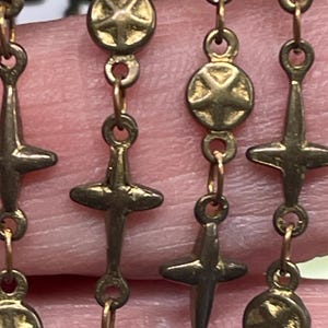 May include: Close-up of a gold-colored rosary chain with cross and star-shaped bead details. The chain is made of small, linked rings. The crosses have a simple, four-arm design.