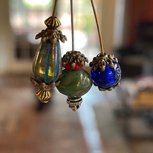 May include: Three decorative hat pins with ornate metalwork and colorful glass beads. The beads are teardrop-shaped, round, and oval, in shades of blue, green, and cobalt. The metalwork has a vintage, antique appearance.