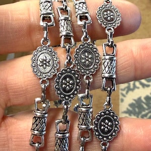 May include: Silver-toned bracelet featuring a series of decorative links. The design includes floral-shaped medallions and textured cylindrical links, connected by rectangular clasps. The bracelet has an antique or vintage aesthetic.