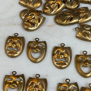 May include: A collection of 17 gold-toned metal charms shaped like theatrical masks. Each mask has a different expression, some are smiling, some are frowning, and some are neutral.