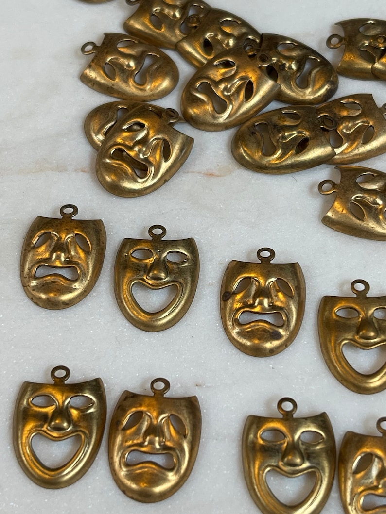 May include: A collection of 17 gold-toned metal charms shaped like theatrical masks. Each mask has a different expression, some are smiling, some are frowning, and some are neutral.