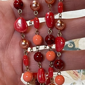 May include: A multi-strand necklace featuring a variety of beads in shades of red, coral, and peach. The beads are connected by gold-colored chains. The beads include round, oval, and irregular shapes.
