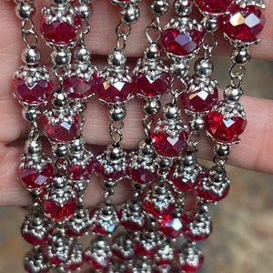 May include: A strand of red crystal beads with silver accents. The beads are faceted and have a shiny finish. The strand is made of multiple rows of beads, creating a layered effect.