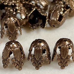 May include: Close-up of several bronze-colored, decorative bead caps. The caps have a floral design with leaf and scroll details. The caps are open at the top and have a small hole for stringing beads. These are ideal for jewelry making.