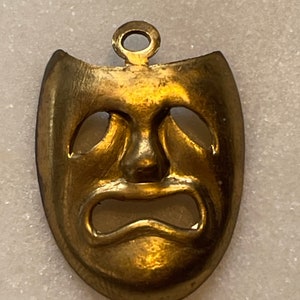 May include: A gold-colored metal pendant in the shape of a tragic mask. The mask has a sad expression with downturned eyes and a wide, open mouth.